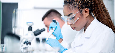 Laboratory technician wearing safety goggles and gloves uses a pipette to handle liquid samples at a lab bench, with another technician working in the background