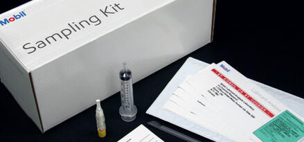 Mobil-branded sampling kit containing a box labeled ‘Sampling Kit,’ a syringe, a small sample vial, a spatula, and several laboratory analysis forms arranged on a black surface