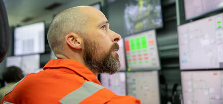 Industrial operator wearing protective clothing monitors data on multiple screens in a control room