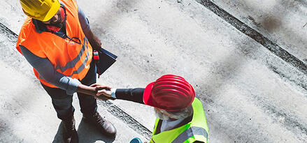 Two construction workers wearing safety gear shaking hands on a jobsite, viewed from above.