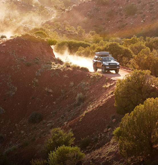A black SUV is driving along a narrow dirt road through a rugged desert landscape
